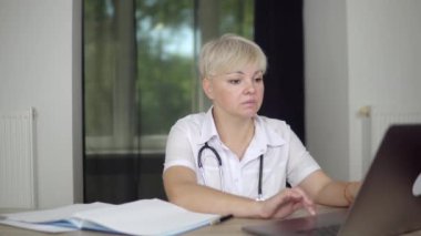 The blonde concentrated women physician is checking the data about patients. Work notebooks on the top f the table. Light spacious cabinet.