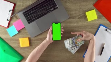 Guy is showing on the phone how easy to earn money with freelance green screen mockup chroma key. Waving american 100 dollar bills. Laptop, color stickers and notebooks.