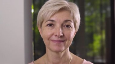 The joyful attractive aged woman with brown eyes and gold chain on the neck with white wall and window on the background.