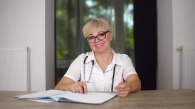 A pleasant women doc with short haircut in glasses, medical gown and stethoscope is giving recommendations about illness to patient at home online video chat.