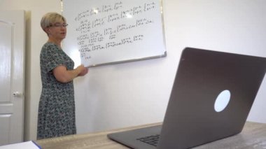 Adult woman teacher in glasses is explaining maths topic to students online in the cabinet in minimalism style with white walls and whiteboard.