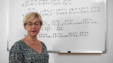 Adult woman teacher in glasses is explaining maths topic to students in the cabinet in minimalism style with white walls and whiteboard.