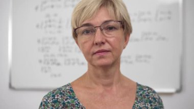 Aged concentrated blonde woman lecturer with poor eyesight in glasses and dress front portrait. Whiteboard with equations and algebra materials on the background.