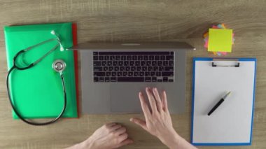 The physician is working with laptop at the cab at clinic. Files, notebook, stethoscope the wooden table.