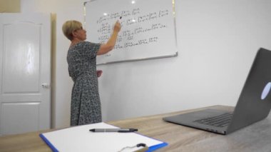 Serious aged mathematics lecturer in dress and glasses is explaining equation topic online on video call to students at office.