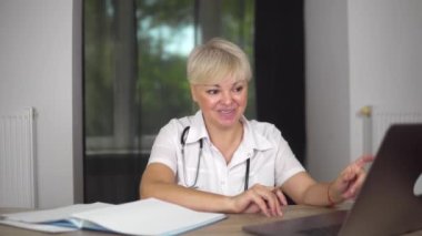 The concentrated blonde women doctor in medical gown with stethoscope is giving the recommendation online to ill patients. Notebook with pen on the table.