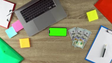 Freelancer is showing on the phone in horizontal position how easy to earn money with freelance green screen mockup. Laptop, color stickers and notebooks.