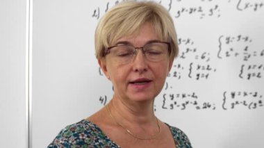 Elderly blonde woman with short haircut maths lecturer is explaining material from whiteboard to her students.