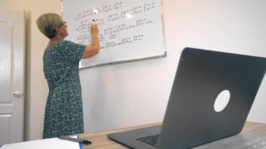 Adult blonde woman maths lecturer is explaining material from whiteboard to students on video chat.