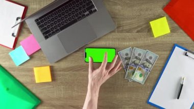 Guy is showing on the phone in horizontal position how easy to earn money with freelance green screen mockup chroma key. 100 dollar bills on the table. Laptop, color stickers and notebooks.