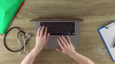 The physician is working at the table on the laptop. Files, notebook, stethoscope on the wooden table.