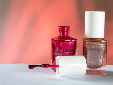 Kırmızı arka planda ve beyaz yüzeyde iki izole tırnak cilası. Bir cilası parlak ve bej. İkinci cila ise açık kapaklı kırmızı parlaklıkta. Yakınlarda bir fırça var.