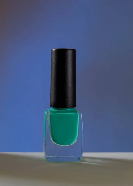 In the foreground, a transparent bottle with turquoise nail polish close-up. The background is blue. Beauty and nail care woman.