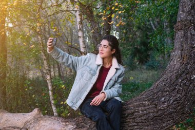 Bir ağaç dalında cep telefonuyla selfie çeken genç bir kadın..