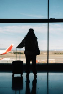 Havaalanının penceresinden dışarı bakan bavullu bir kadının silueti. Seyahat kavramı.