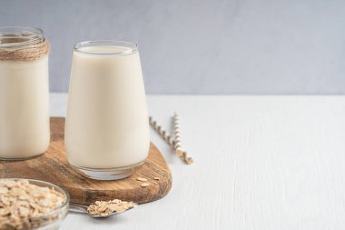 Sağlıklı vejetaryen bitkisel yulaf sütü kremalı yulaf ezmesi aromalı ve tahta tahtada içilen cam bardakta servis edilen ve kaşıkla beyaz masadaki malzemelerle dolu. Kopyalama alanı olan resim 