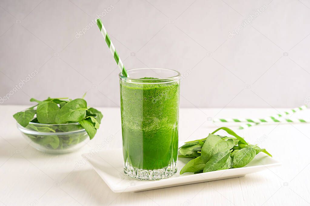 Green vegetarian healthy homemade spinach smoothie or juice served in drinking glass with paper straw and leaves ingredient on plate and bowl on white wooden table. Dieting and weight loss concept