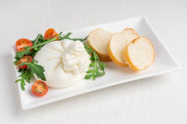 Ev yapımı organik burrata mozzarella ve kremadan yapılmış İtalyan sütlü peyniri kiraz domatesli, roka yeşil yapraklı ve beyaz ahşap zemin üzerinde dilimlenmiş ekmek servis edilir.