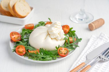Burrata İtalyan sütü mozzarella ve kremadan yapılmış yumuşak organik peynir. Çeri domatesli, roka yeşili yapraklı, dilimlenmiş ekmek, sofra takımı ve beyaz ahşap masada bir bardak şarapla servis ediliyor.