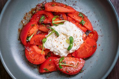 Ortasında yumuşak beyaz burrata İtalyan inek sütü peyniri olan taze, sulu kırmızı domateslerin üst görüntüsü. Akşam yemeğinde restoranda gri tabakta servis edilen mikroyeşil ve biber baharatlarıyla süslenmiş.
