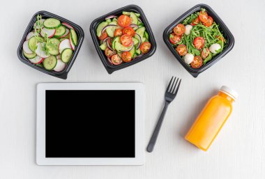 Salatalık dilimleri, roka ve domateslerden oluşan çeşitli vejetaryen salatalar portakal suyu, çatal ve tablet bilgisayarlı siyah tek kullanımlık yemek kaplarında servis edilir.