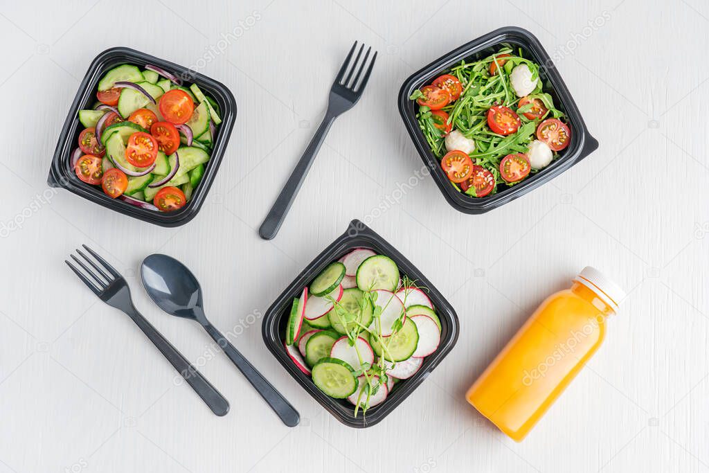 Variety of vegan vegetable salads made of cucumber slices, arugula and tomatoes served in black disposable lunch containers or boxes with orange juice bottle and tableware on white wooden background