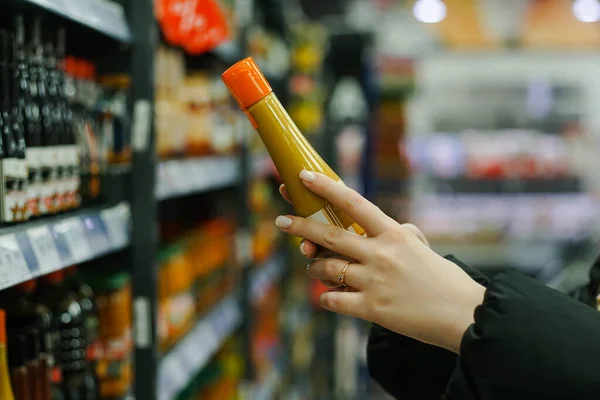 Female hand choosing yellow mustard sauce plastic bottle at store, close up — стоковое фото
