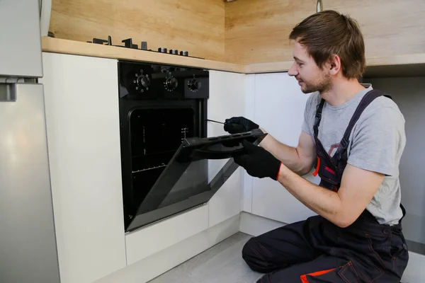 Young man worker in uniform repairing electric oven in kitchen.
