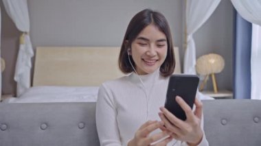 Mutlu genç bir kadın kulaklık takıyor. İnternetten görüntülü konuşma yapıyor. Telefona bakıyor, kanepeye oturuyor, gülümsüyor..