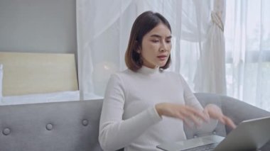 Stresli genç bir kadın el ele tutuşup dizüstü bilgisayara bakıyor, projeyle ilgili sorunları var, oturma odasında kanepede oturuyor..
