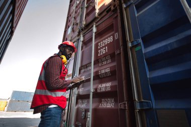 Afro-amerikan erkek işçi elinde tablet tutuyor ve kargo nakliye gemisinden gelen yük konteynırlarını kontrol ediyor..