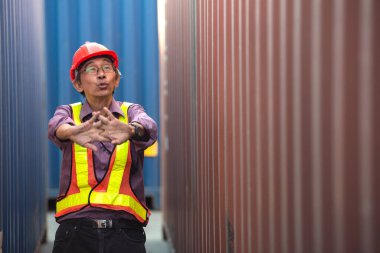 Güvenlik yeleği ve miğfer giyen yaşlı Asyalı işçi mühendisi, el kaldırıyor, lojistik kargo konteynırlarının arasında duruyor..