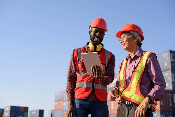 Happy African American and senior asian male worker using tablet during video call in a logistic shipping cargo containers yard.