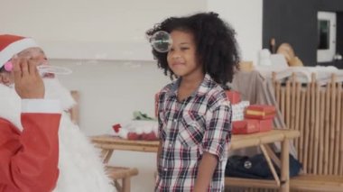Noel Baba elbisesi giyen bir adam oturma odasında kız çocuğuyla oynayan baloncuklar çıkarıyor..