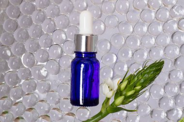 Mavi camdan yapılmış kozmetik şişesi, üst görünüm. Etraflarında şeffaf hidrojel topları. Çerçevesi beyaz çiçekli yeşil dallar..
