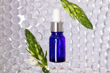 Mavi camdan yapılmış kozmetik şişesi, üst görünüm. Etraflarında şeffaf hidrojel topları. Çerçevesi beyaz çiçekli yeşil dallar..