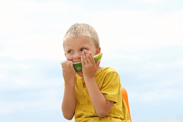 Funny sun tanned blonde kid on the coast, holding juicy slice of the watermelon and eating it with gusto.Summertime concept. — Stock Photo, Image