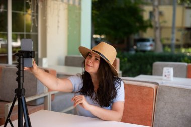 Attractive smiling woman outside sitting with tripod and smartphone,making video call or providing online lesson.