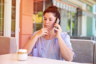 Dışarıda akıllı telefonlu ciddi bir kadın, bir şeyler düşünüyor, arka planda güneş ışığı..