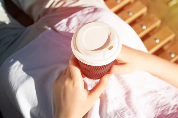 Photography from above of anonymous woman, holding craft paper cup with coffee outside,sunlight on background.