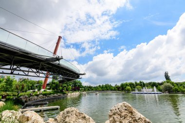 Romanya 'nın Bükreş kentindeki Moghioros Park olarak da bilinen Drumul Taberei Parkı' ndaki çağdaş kırmızı metalik köprü, göl ve canlı yeşil ağaçlara sahip manzara.