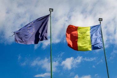 Avrupa Birliği ve Rumen ulusal bayrakları güneşli bir bahar gününde bulutlu mavi gökyüzüne doğru rüzgarda dalgalanıyor