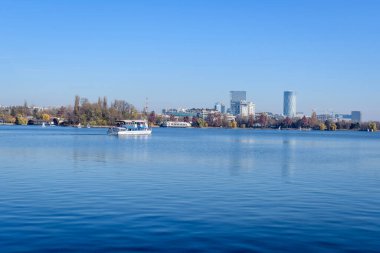 Bucharest, Romania, 21 November 2021: Landscape with white boat on Herastrau lake and large green trees in King Michael I Park (Herastrau) in Bucharest, Romania, in a sunny autumn day