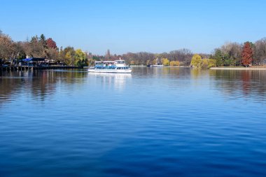 Bucharest, Romania, 21 November 2021: Landscape with white boat on Herastrau lake and large green trees in King Michael I Park (Herastrau) in Bucharest, Romania, in a sunny autumn day