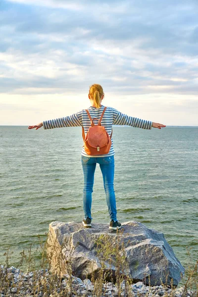 A girl, in pants and a sweatshirt, with a backpack on her back, arms outstretched, standing on a large rock and looking at the sky and water in front of her, rear view.