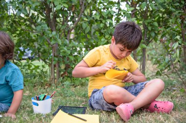 İki öğrenci sırt çantalarıyla parkta yatıyor, biri deftere yazıyor.