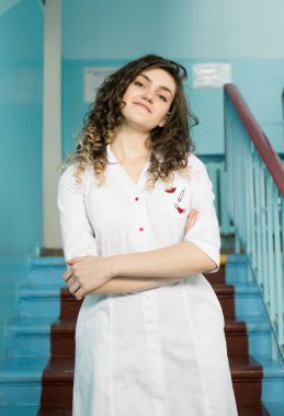 Beyaz önlüklü genç bir kadın doktor bir hastanenin arka planında duruyor.