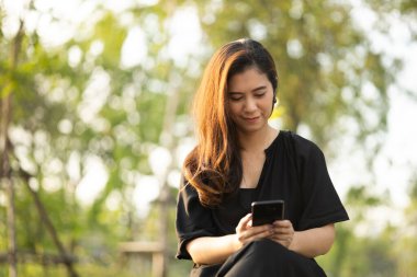Asian woman mature using cell phone in public park. Adult female holding cell phone with beautiful sun light.