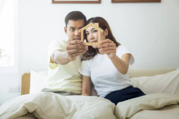 Couple or 2 asian people on bed in bedroom, home or house. Man and woman hold home model in hand together. Look happy, romantic. Concept for married, family, rent, buy, real estate and propertry.