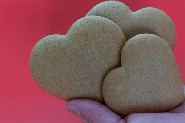 three heart shaped cookies are held by an unrecognizable human hand on a red background. Valentines Day, greeting card, pastry, copy space, love concept, engagement, copy space, horizontal